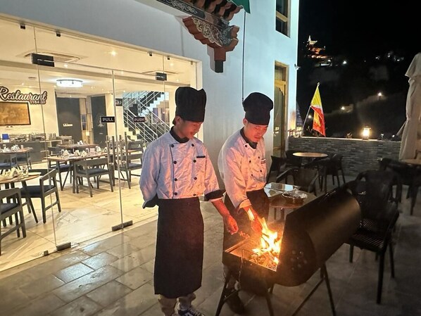 Zwei Köche bereiten draußen bei Nacht Speisen zu, im Hintergrund ist ein Restaurant zu sehen.