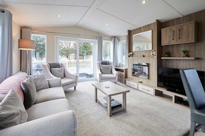 Ein modernes Wohnzimmer mit einer Couch, Sesseln, einem Couchtisch und einem Fernseher. Ein Kamin, ein großes Fenster und ein Balkon mit Vorhängen sind ebenfalls vorhanden.