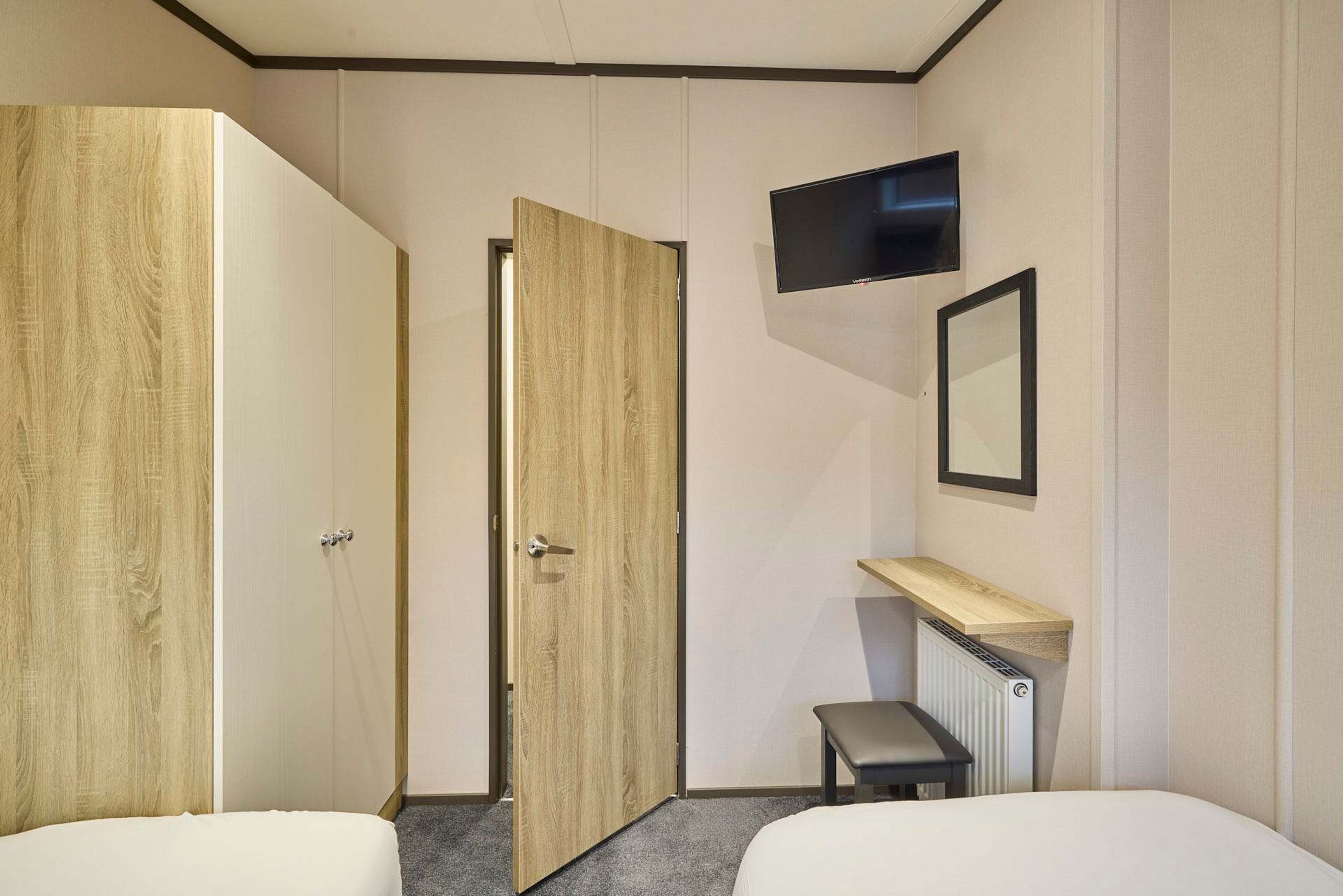 A hotel room with two beds, a wooden wardrobe, a TV mounted on the wall, and a small bench.