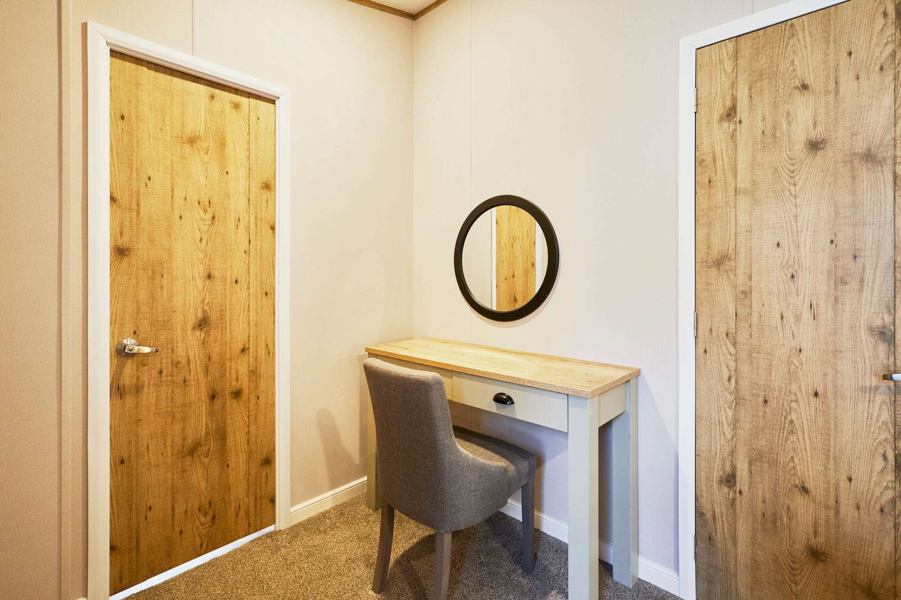 A corner room with a wooden door, a desk with a chair, and a round mirror on the wall.
