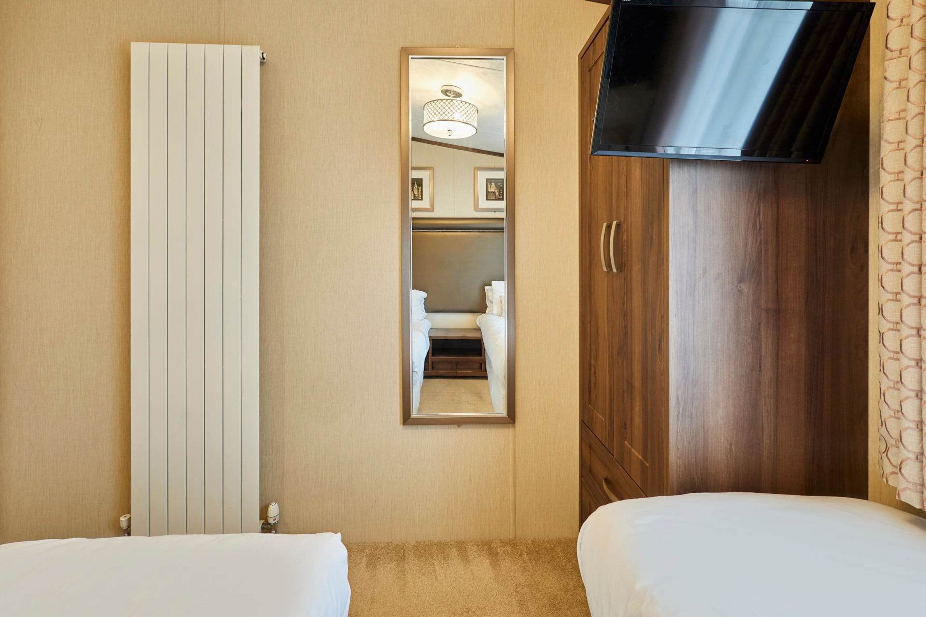 Een hotelkamer met twee bedden, een houten garderobe, een televisie en een spiegel die een badkamer met toilet en wastafel weerspiegelt.