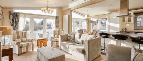 Ein geräumiges Wohnzimmer mit einer großen Couch, Sesseln und einem Couchtisch. Mehrere Fenster sorgen für natürliches Licht und einen Blick ins Freie.