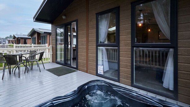 A wooden building with a hot tub on the deck, a covered patio with chairs, and large windows with curtains. - Host Stay Truemans Lodge (York)