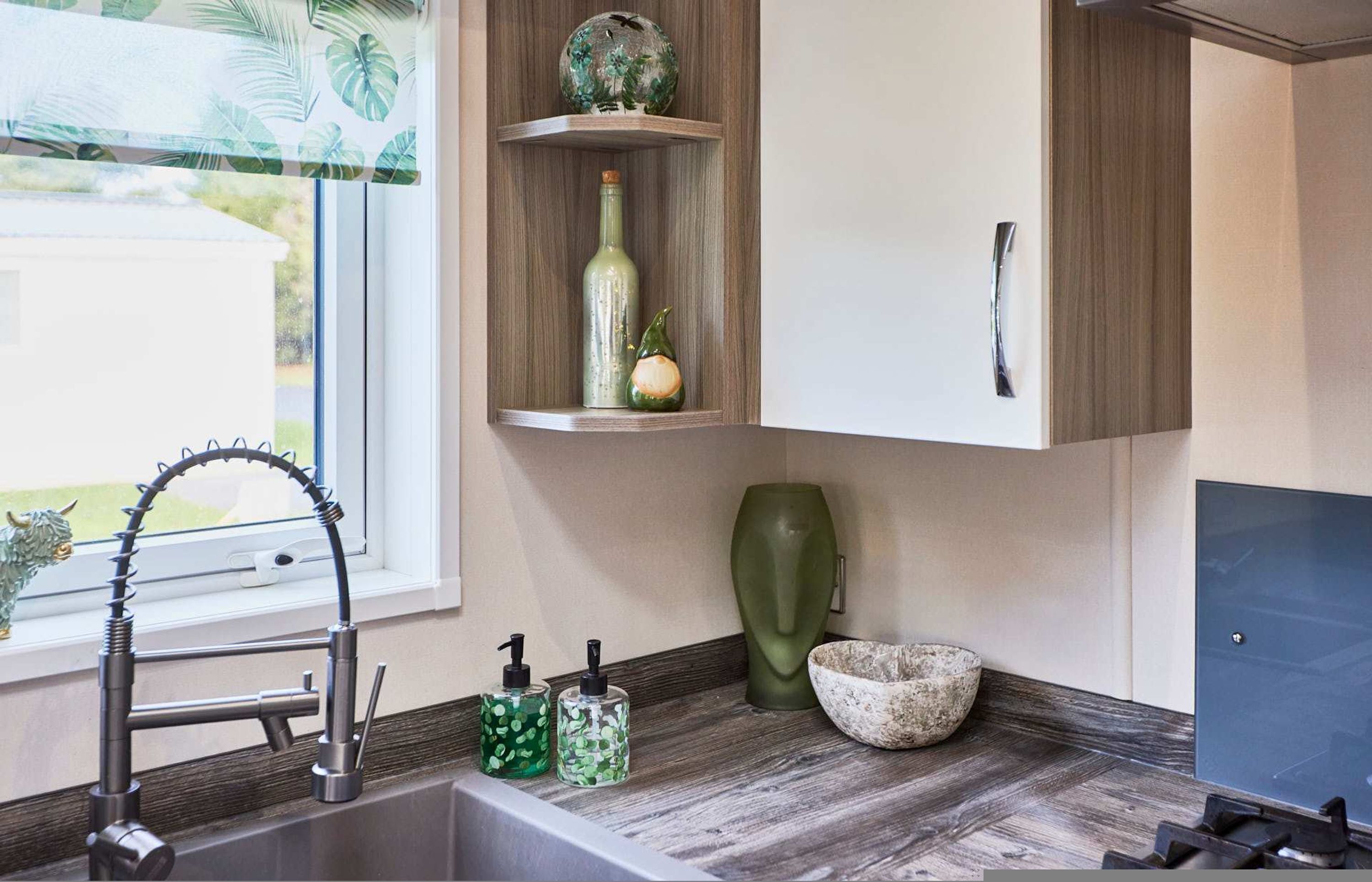 A modern kitchen with a wooden countertop, a built-in shelf with decorative items, a sink with a modern faucet, and a window with a view of greenery.