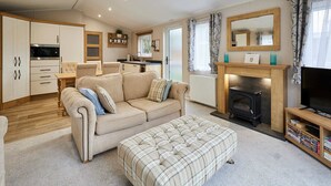 Ein gemütliches Wohnzimmer mit Sofa, Hocker, Kamin, TV-Schrank und einer Küchenzeile mit Schränken und Herd.