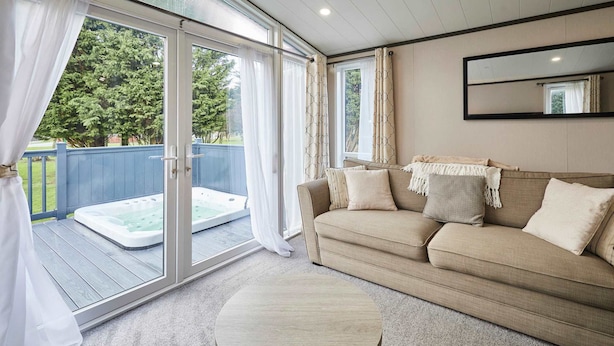 A room with a sliding glass door leading to a deck with a hot tub, a beige sofa with cushions, and a round coffee table. - Host Stay The Kestrel Lodge (York)