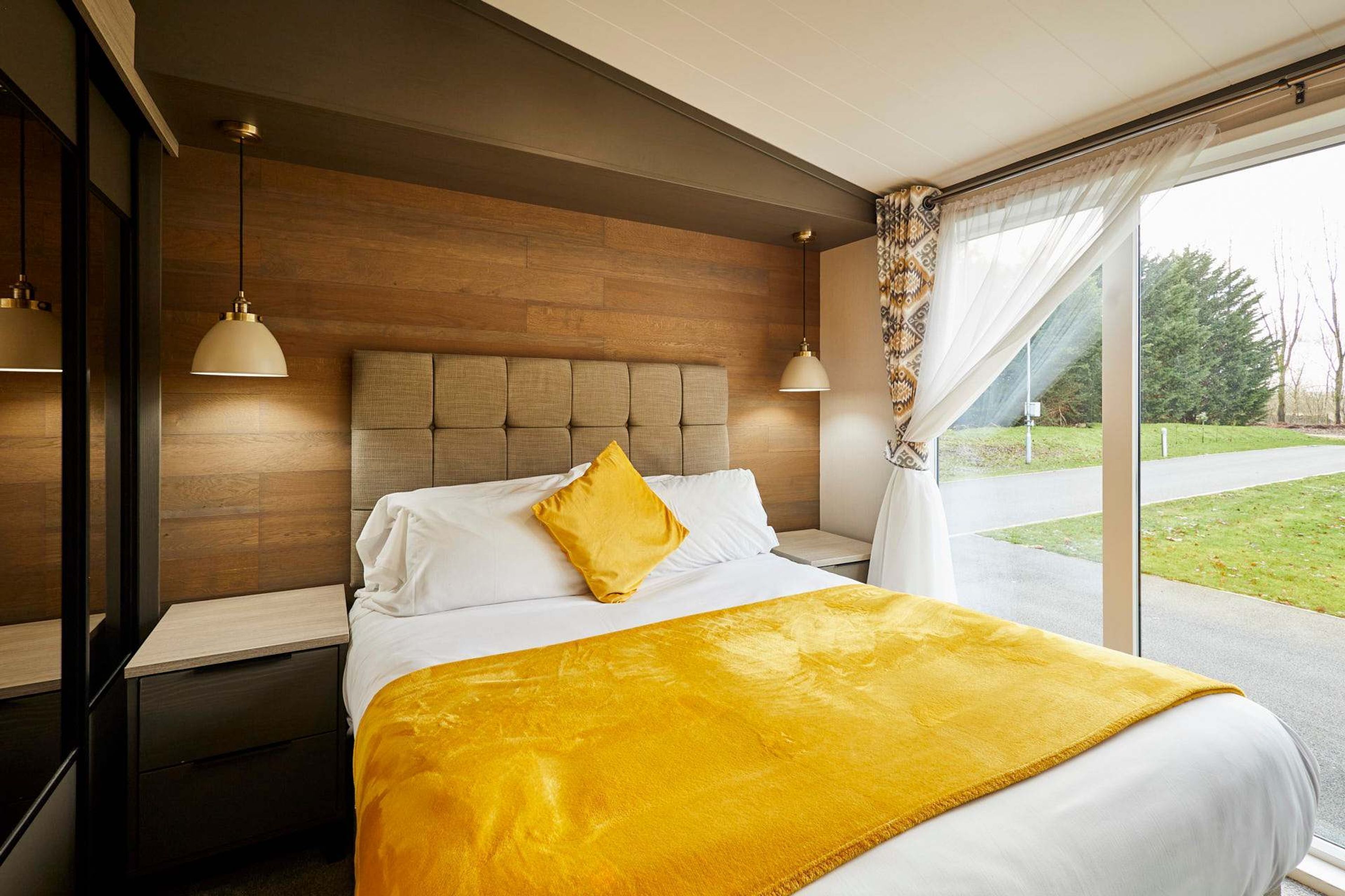A modern bedroom with a wooden headboard, a large bed with a yellow throw blanket, and a view of a green landscape through a large window.