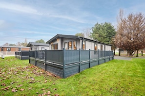 A single-story mobile home with a fenced yard, a grassy area with scattered leaves, and a clear sky. - Host Stay Copse View Lodge (York)