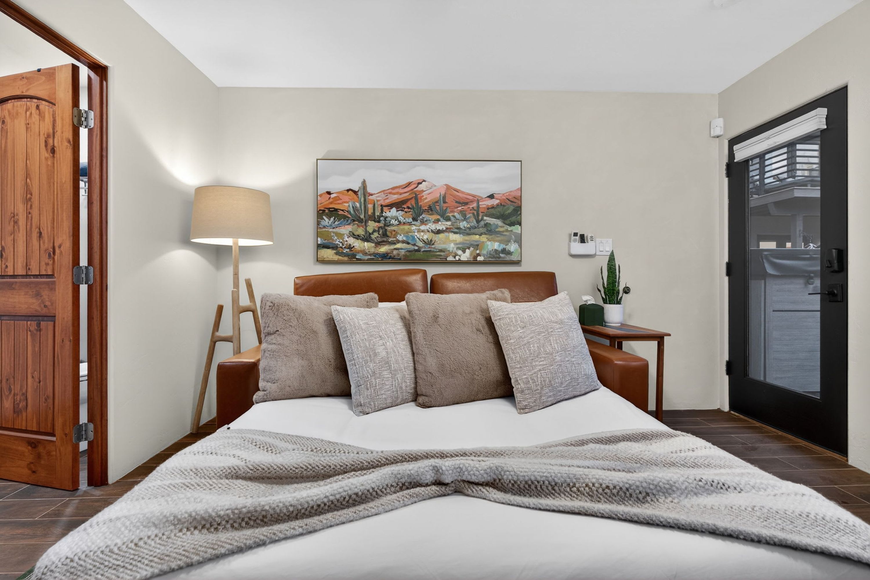 Un dormitorio ordenado con cama, almohadas, una lámpara de noche, un cuadro y una pequeña mesa con una planta.