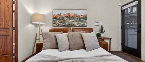 Un dormitorio ordenado con cama, almohadas, una lámpara de noche, un cuadro y una pequeña mesa con una planta.
