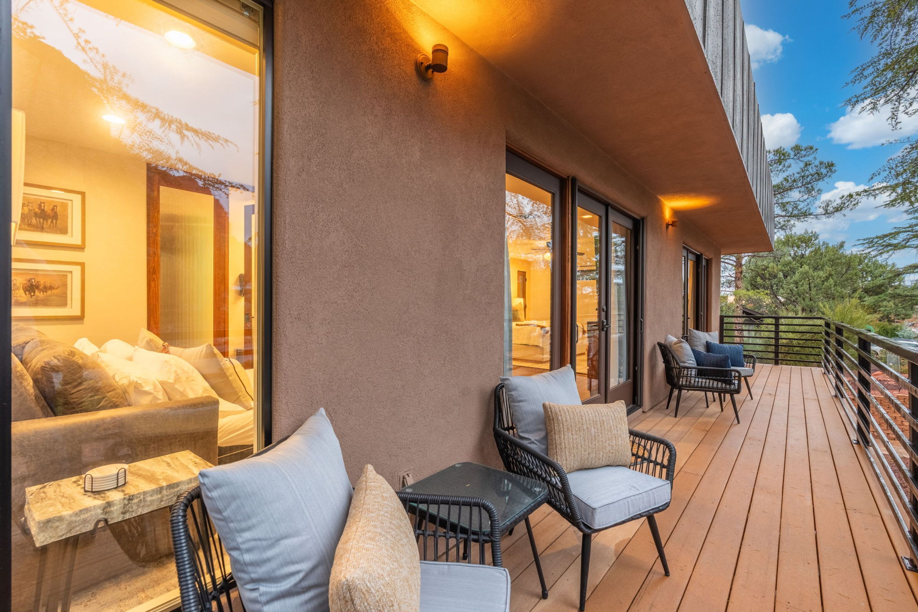 A wooden deck with outdoor seating, including a sofa and chairs, and a glass door leading to an interior space with a bed and framed pictures.