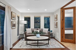 Un salon moderne avec un canapé marron, une table basse ronde et un lampadaire. Les murs sont ornés de tableaux encadrés et une grande fenêtre offre une vue sur les arbres.