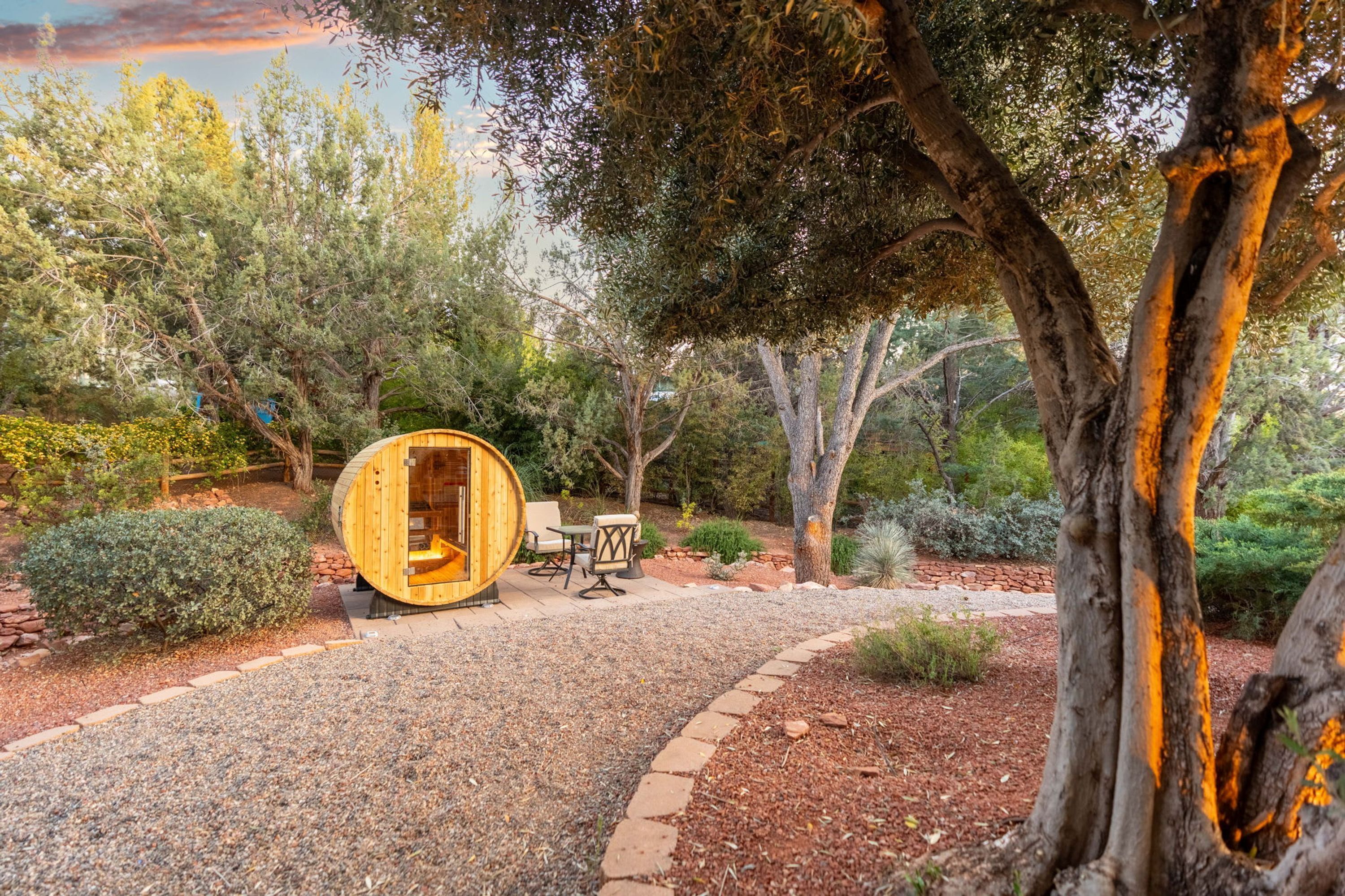 Un área de estar al aire libre con madera, una estructura circular de madera, rodeada de árboles y un camino de grava.