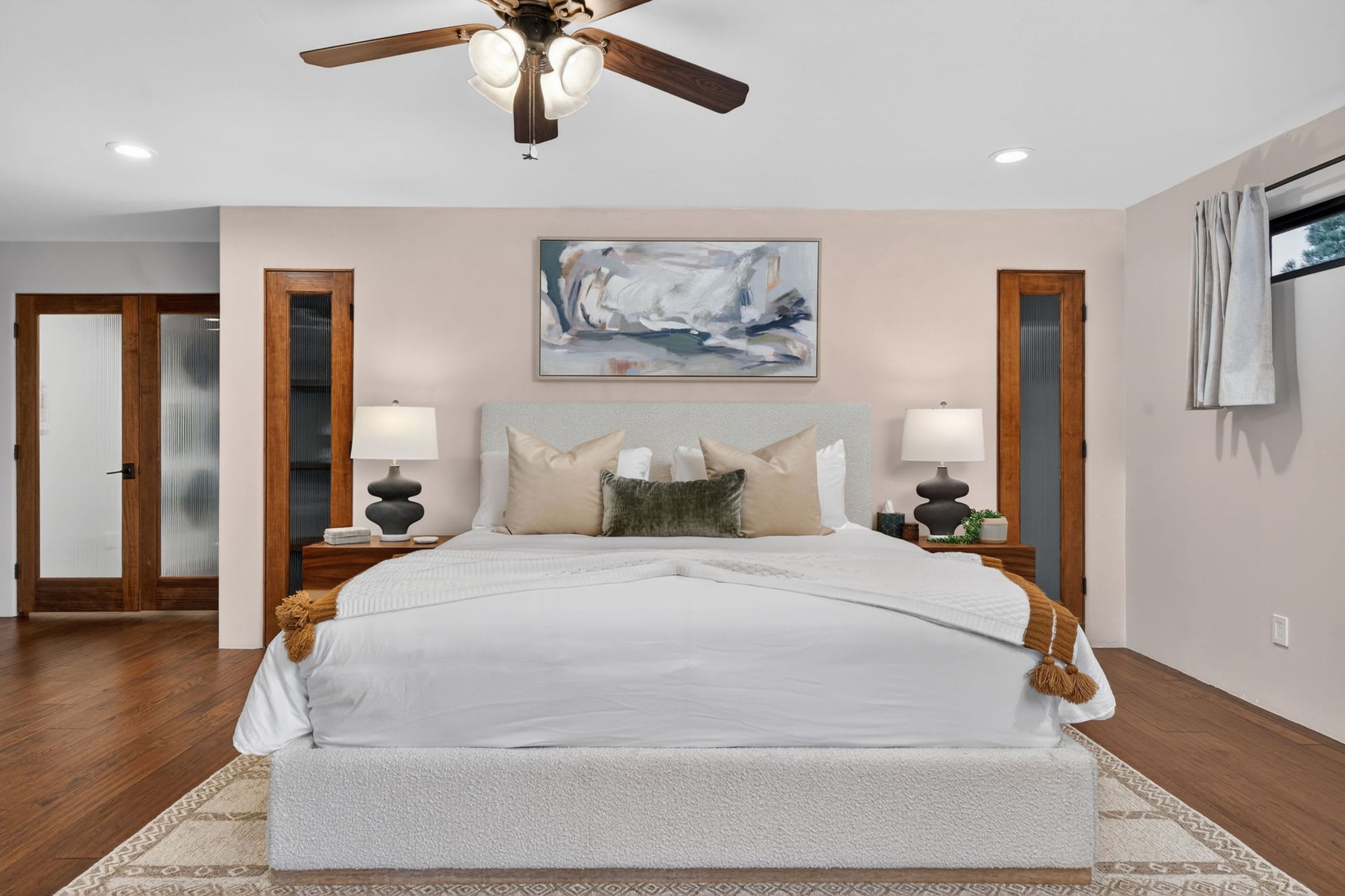 A bedroom with a large bed, two bedside tables with lamps, a ceiling fan, and a painting on the wall.