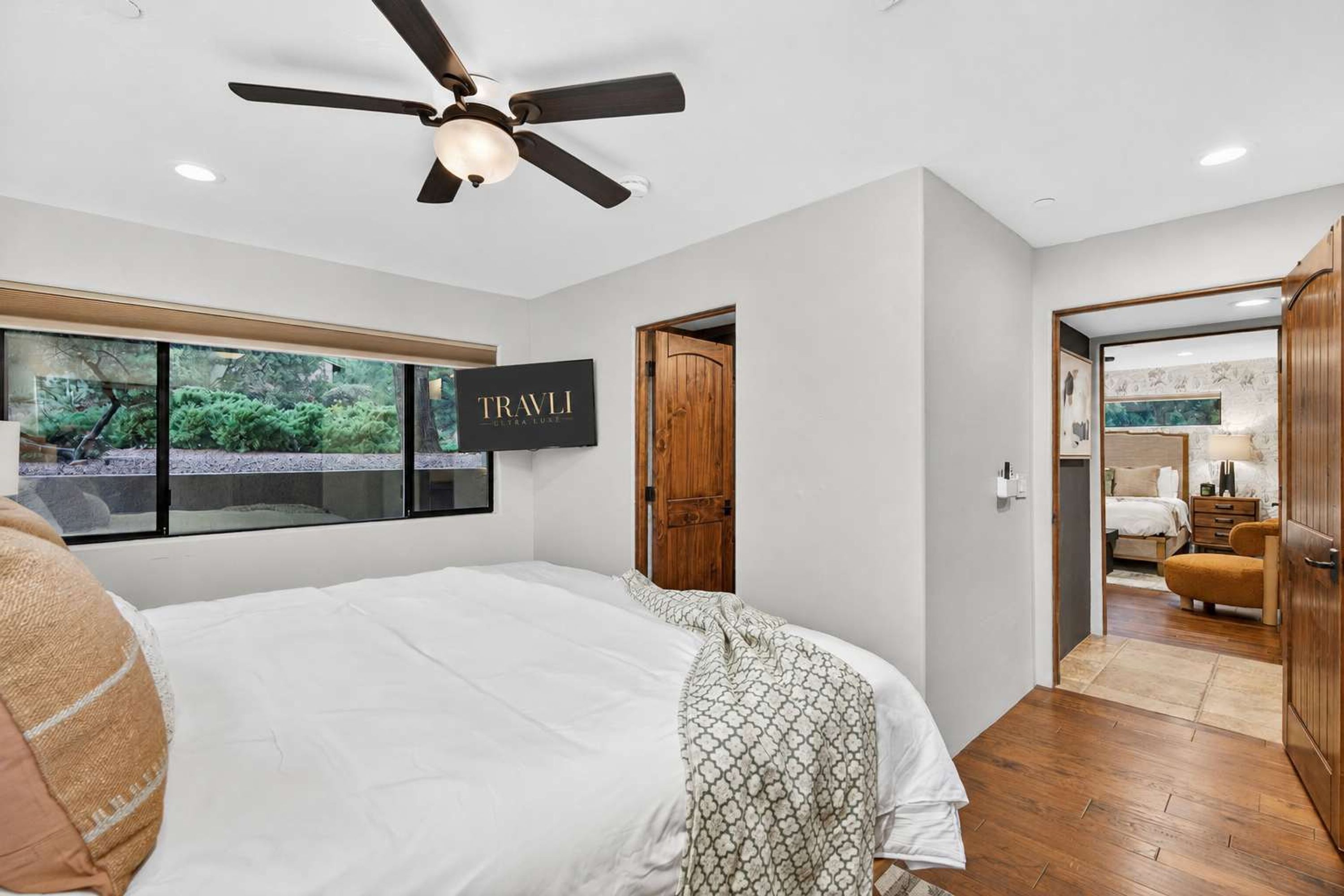 A bedroom with a large bed, a ceiling fan, a window with a view, and a door leading to another room.