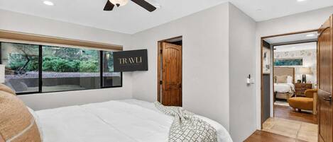 A bedroom with a large bed, a ceiling fan, a window with a view, and a door leading to another room.