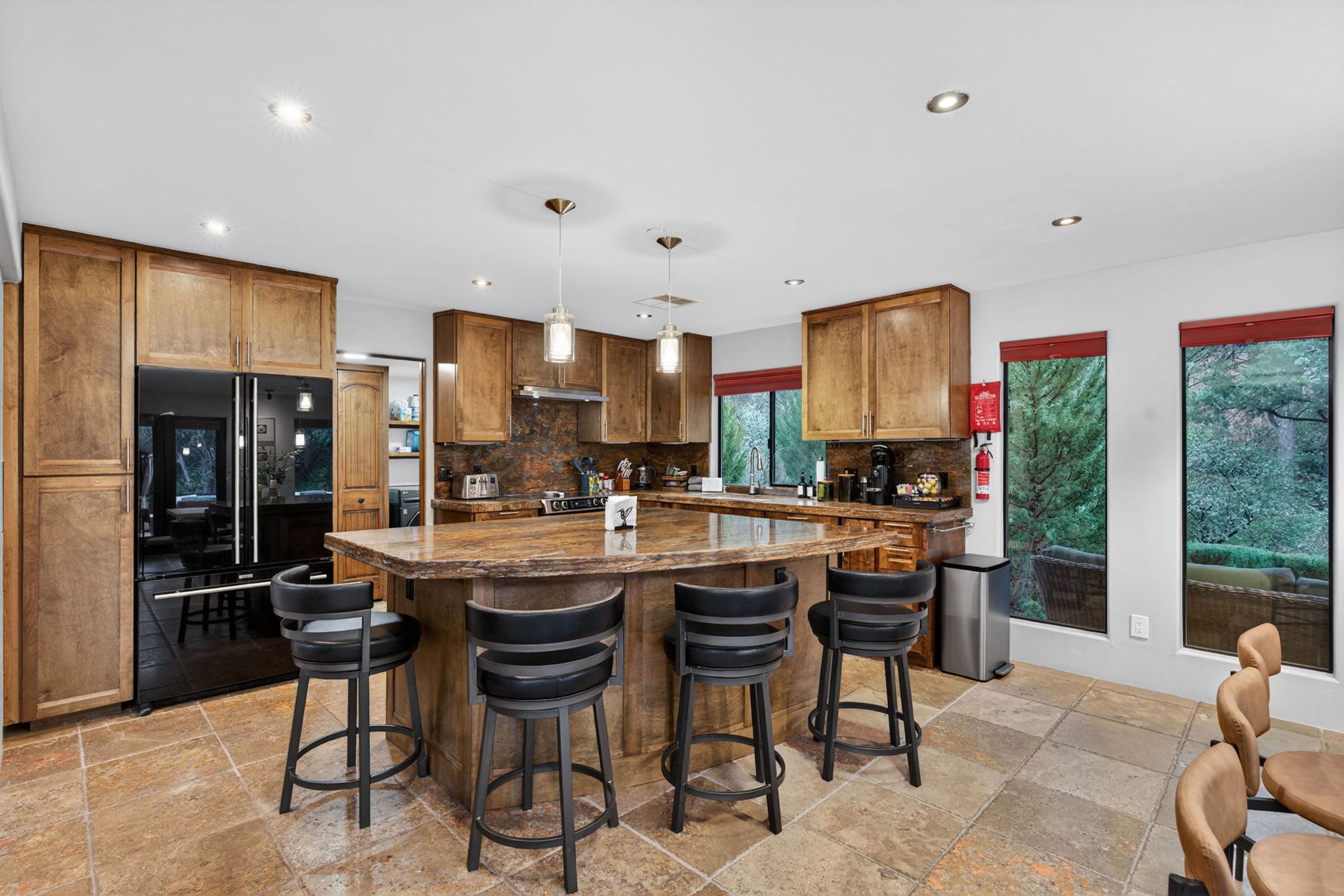 A modern kitchen with wooden cabinets, a central island with stools, and large windows offering views of greenery.
