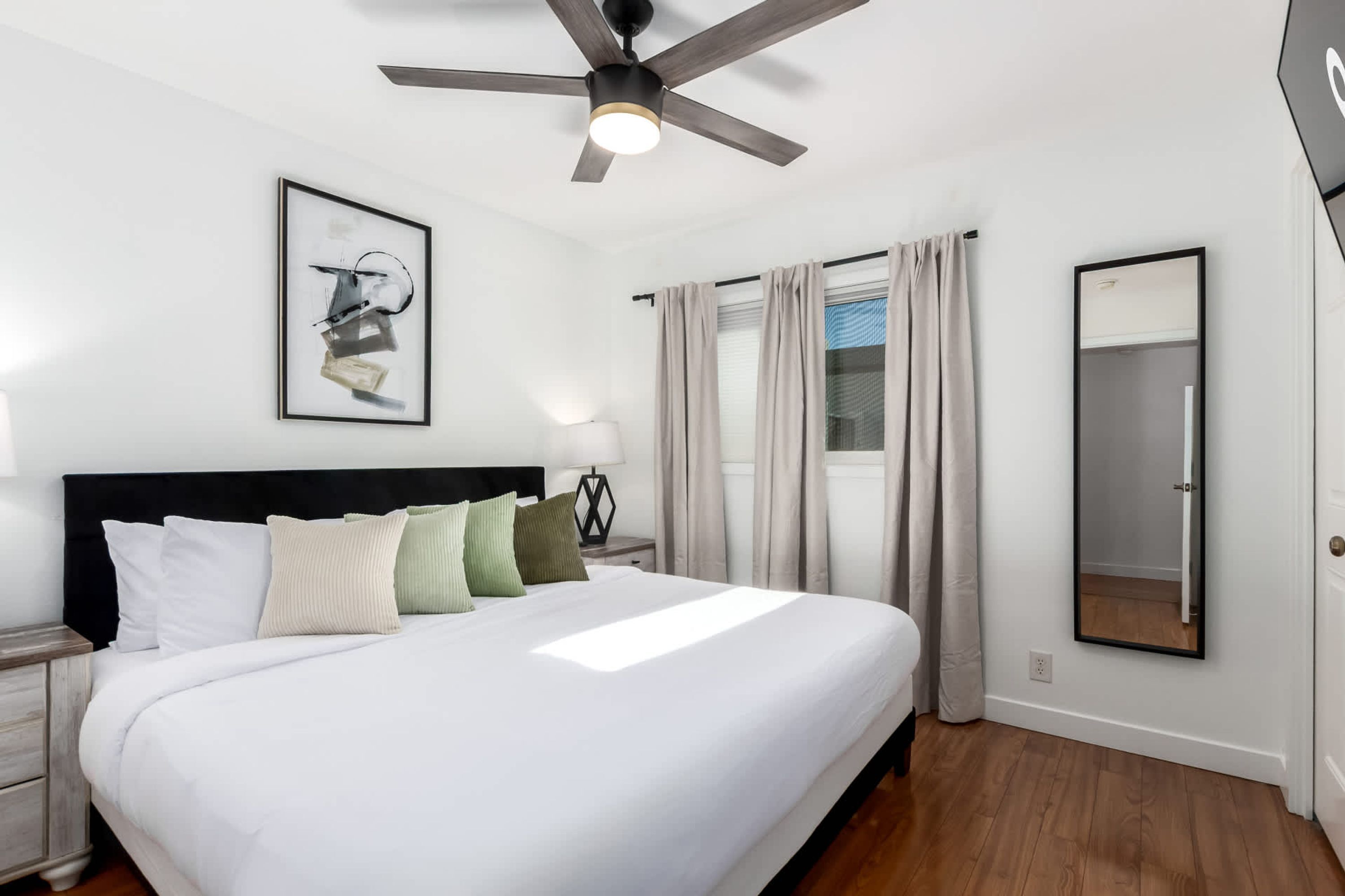 A modern bedroom with a large bed, a ceiling fan, a framed abstract artwork, a bedside table with a lamp, and a mirror on the wall.