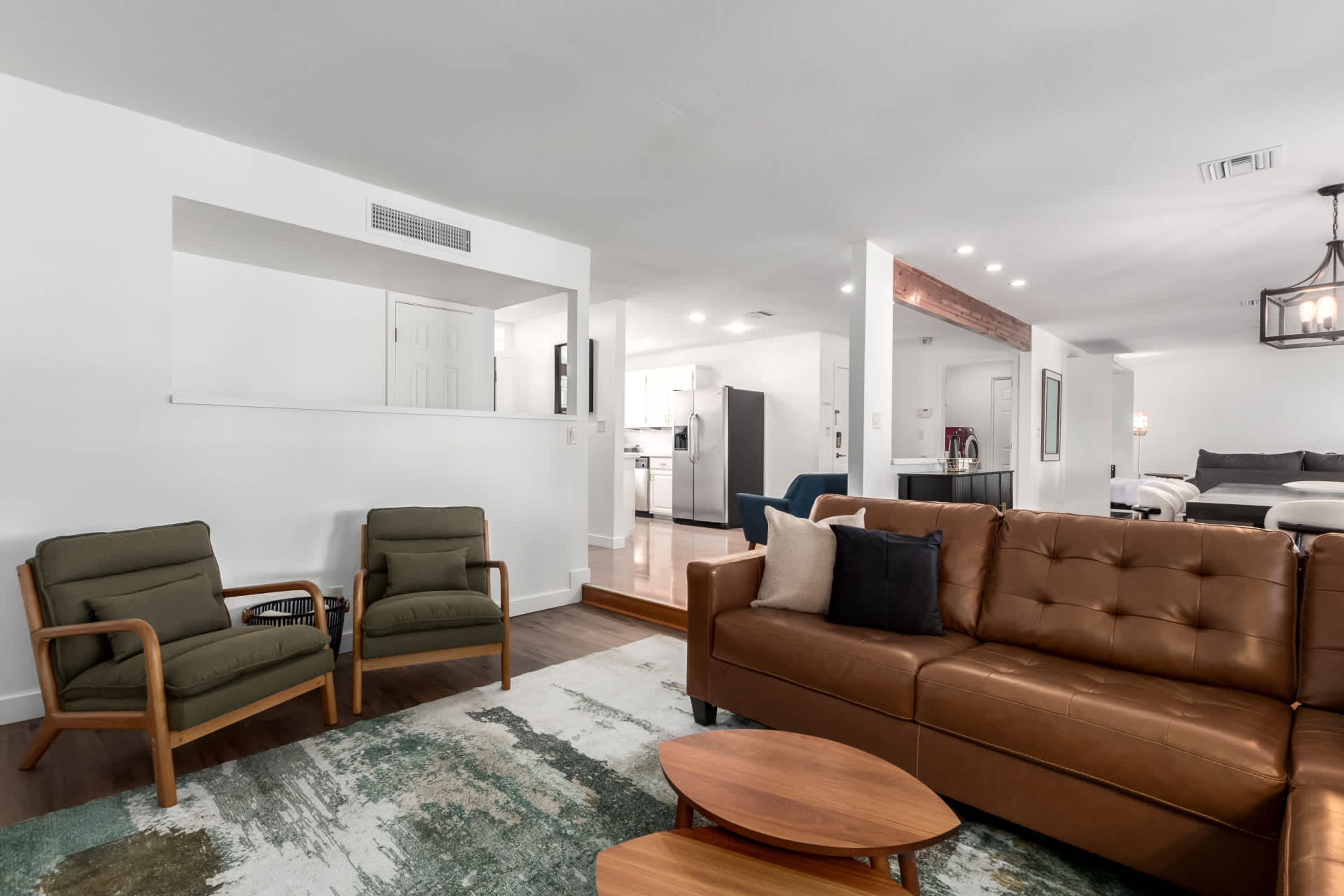 A modern living room with a brown leather sofa, two green armchairs, and a wooden coffee table. A kitchen is visible in the background.
