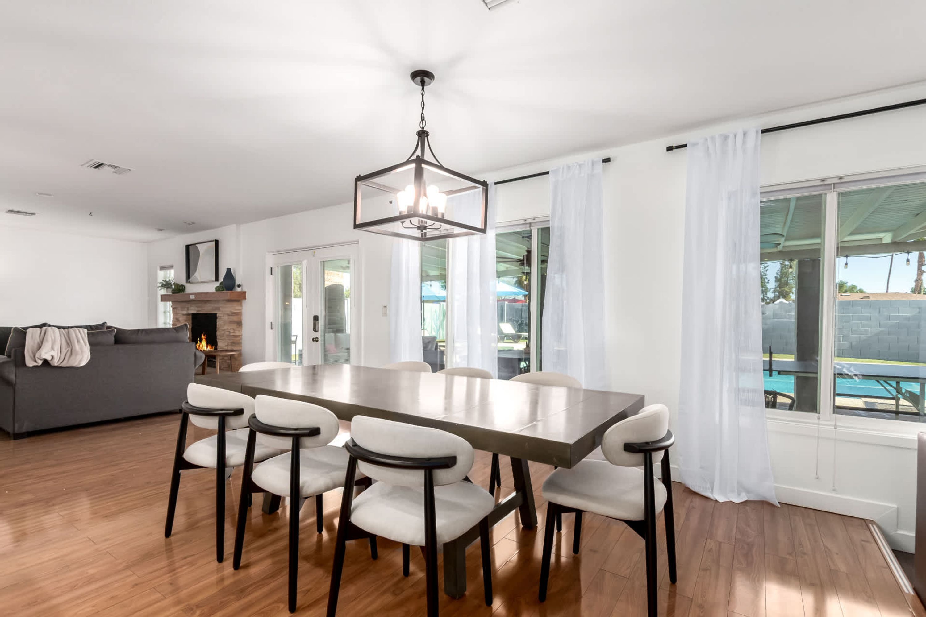 Un'area pranzo moderna con un tavolo grande e sei sedie, un lampadario e vista su una piscina attraverso ampie finestre.