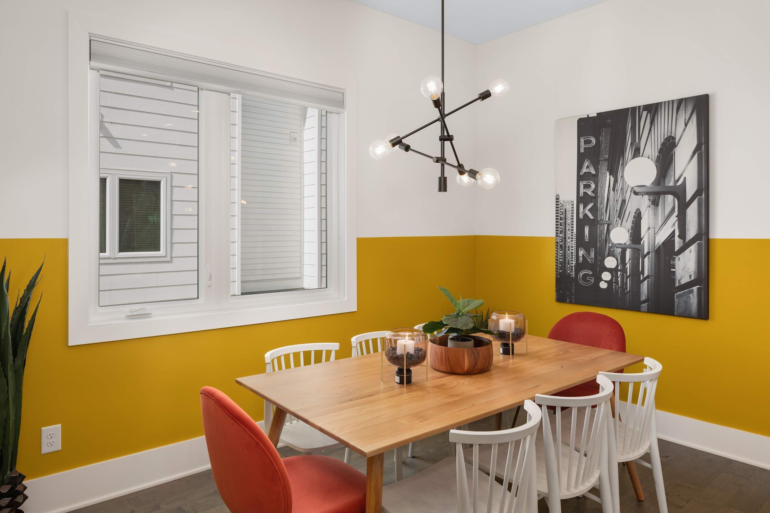 Un coin repas avec une table en bois, des chaises blanches, une chaise rouge, un luminaire suspendu et un tableau accroché au mur.