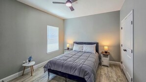 Un dormitorio con cama, ventilador de techo, lámparas de noche, una mesita con un florero y una puerta.