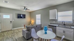 Una sala moderna con un sofá gris, una mesa de comedor blanca con cuatro sillas, un jarrón azul, un ventilador de techo, un televisor de pantalla plana y una zona de cocina con un fregadero y armarios.