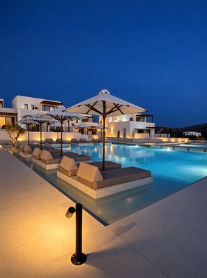 A modern pool area with lounge chairs and umbrellas, surrounded by white buildings with multiple levels. - Agali Hotel Milos (Milos)