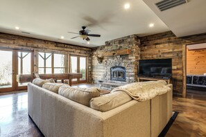 A living room with a stone fireplace, a large sofa, a television, and a ceiling fan. - Game Room + Deck: Spacious Spruce Pine Retreat! (Spruce Pine)