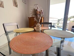 Ein Esstisch mit Glastischplatte und gewebten Platzsets, eine Vase mit getrockneten Blumen und Blick auf einen Balkon mit Geländer.
