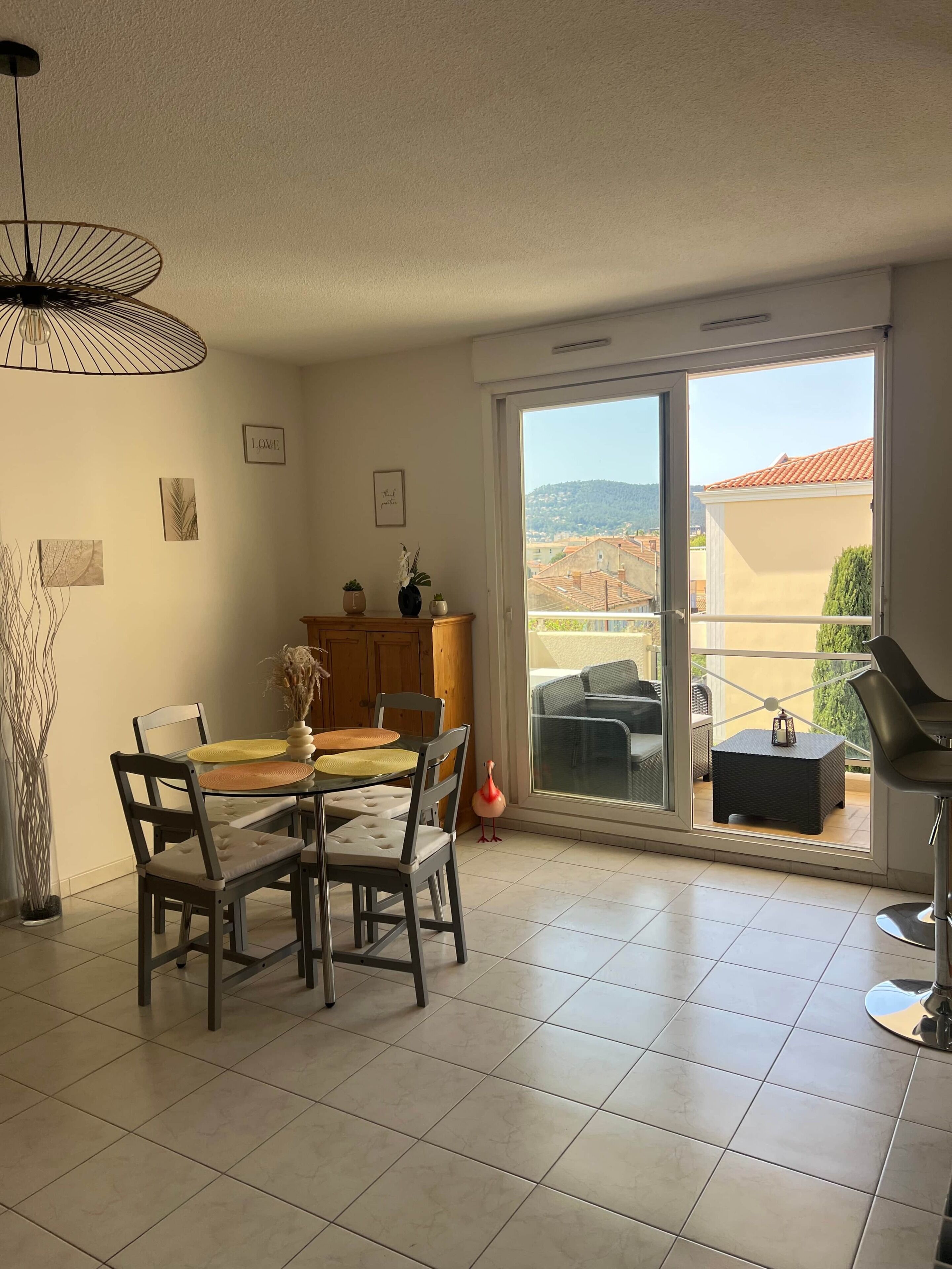 Un comedor con una mesa, sillas, una lámpara colgante y vistas a un área de estar exterior a través de una puerta corrediza de vidrio.