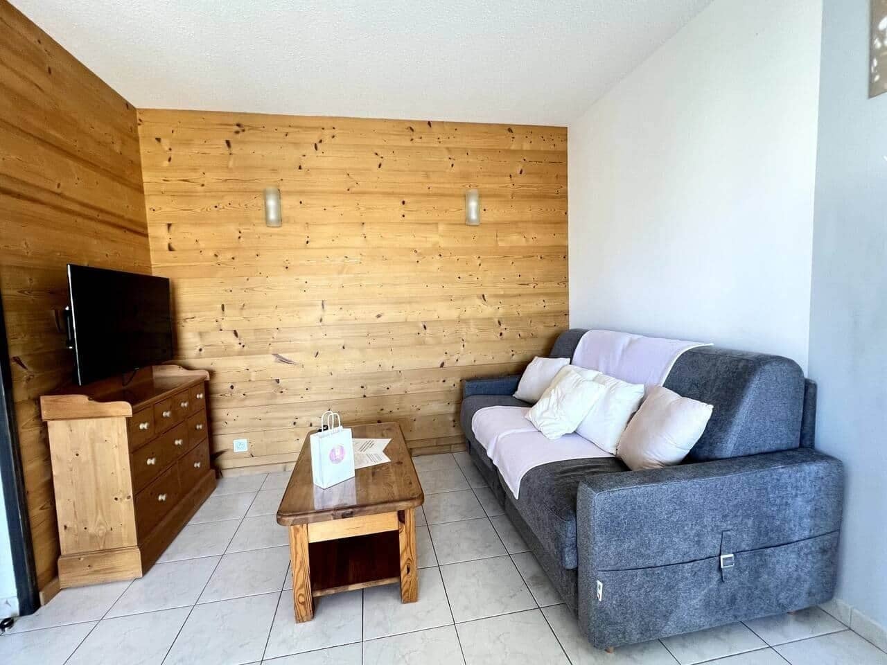 A cozy living room with a wooden wall, a grey sofa, a wooden coffee table, and a TV on a wooden cabinet.