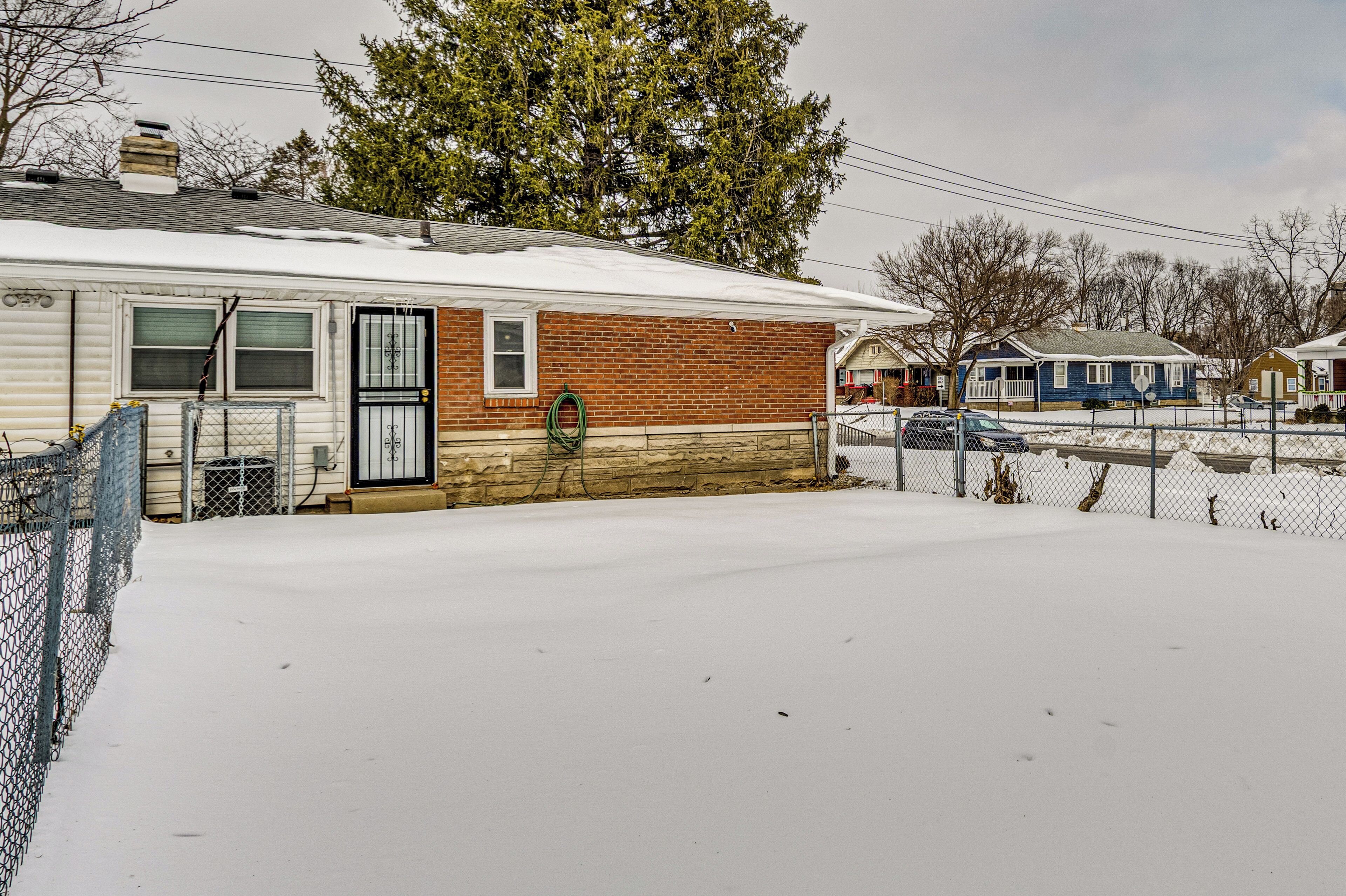 A single-story house with a brick section and a white section, surrounded by a snow-covered yard and a chain-link fence.