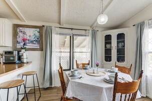 Un comedor con una mesa dispuesta para cuatro personas, sillas de madera, mantel blanco y un cuadro en la pared.