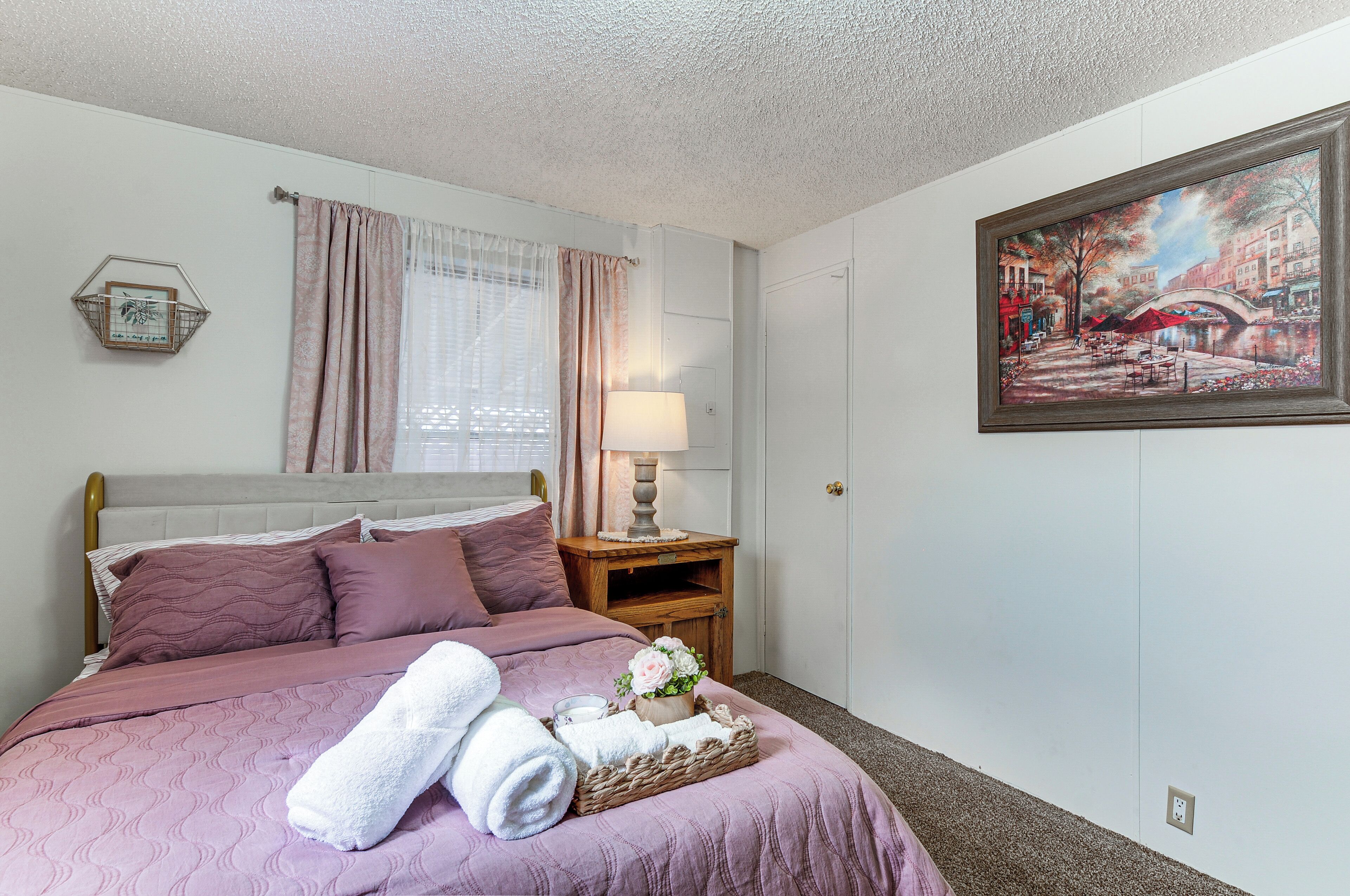 A neatly made bed with pink bedding, a bedside table with a lamp, and a painting of a cityscape on the wall.