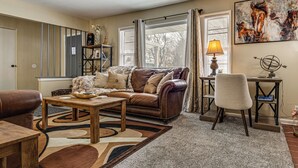 Ein Wohnzimmer mit braunem Ledersofa, einem Holztisch und einem gemusterten Teppich. Eine Lampe steht auf einem Beistelltisch, ein Gemälde hängt an der Wand und ein Fenster mit Jalousien ist vorhanden.