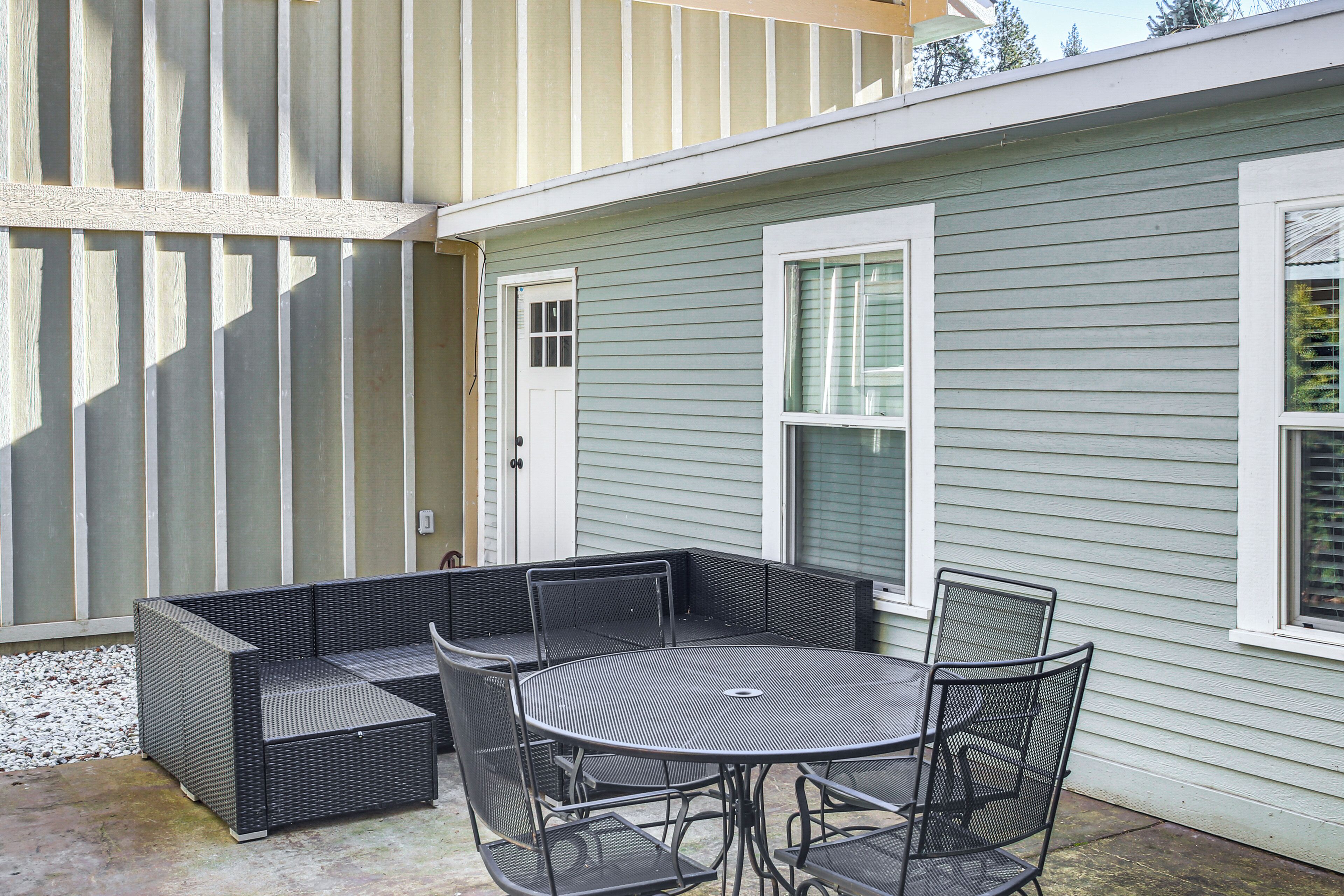 A patio with a black wicker sofa and a round table with chairs, adjacent to a house with light green siding and white trim.
