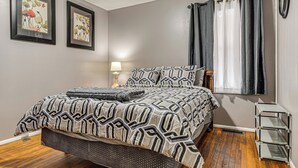 Un dormitorio con una cama, dos cuadros enmarcados en la pared, una lámpara de noche y una ventana con cortinas.