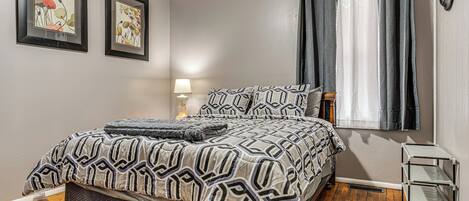 A bedroom with a bed, two framed artworks on the wall, a bedside lamp, and a window with curtains.