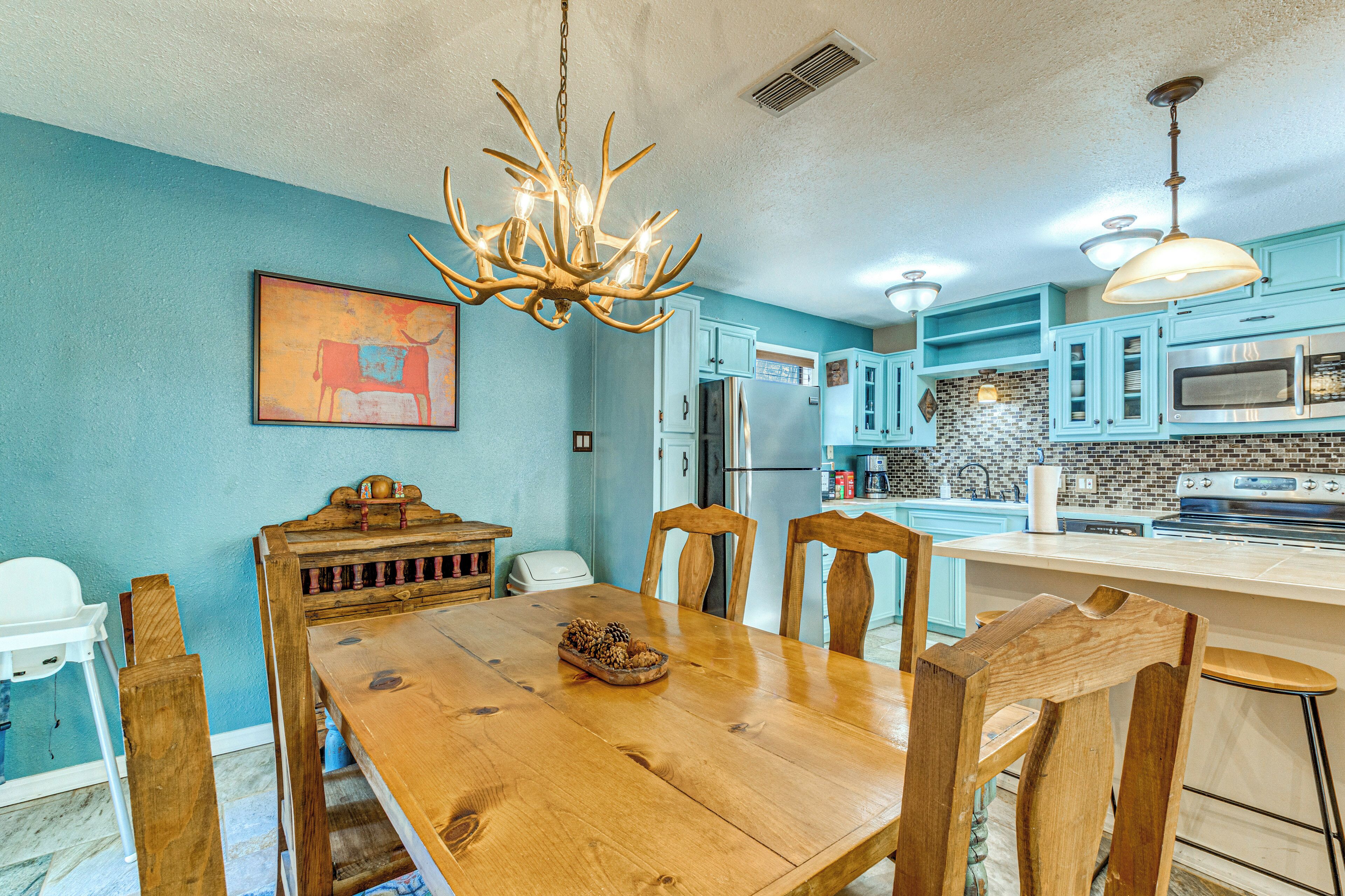 A dining area with a wooden table and chairs, a chandelier, and a kitchen with blue cabinets and stainless steel appliances.