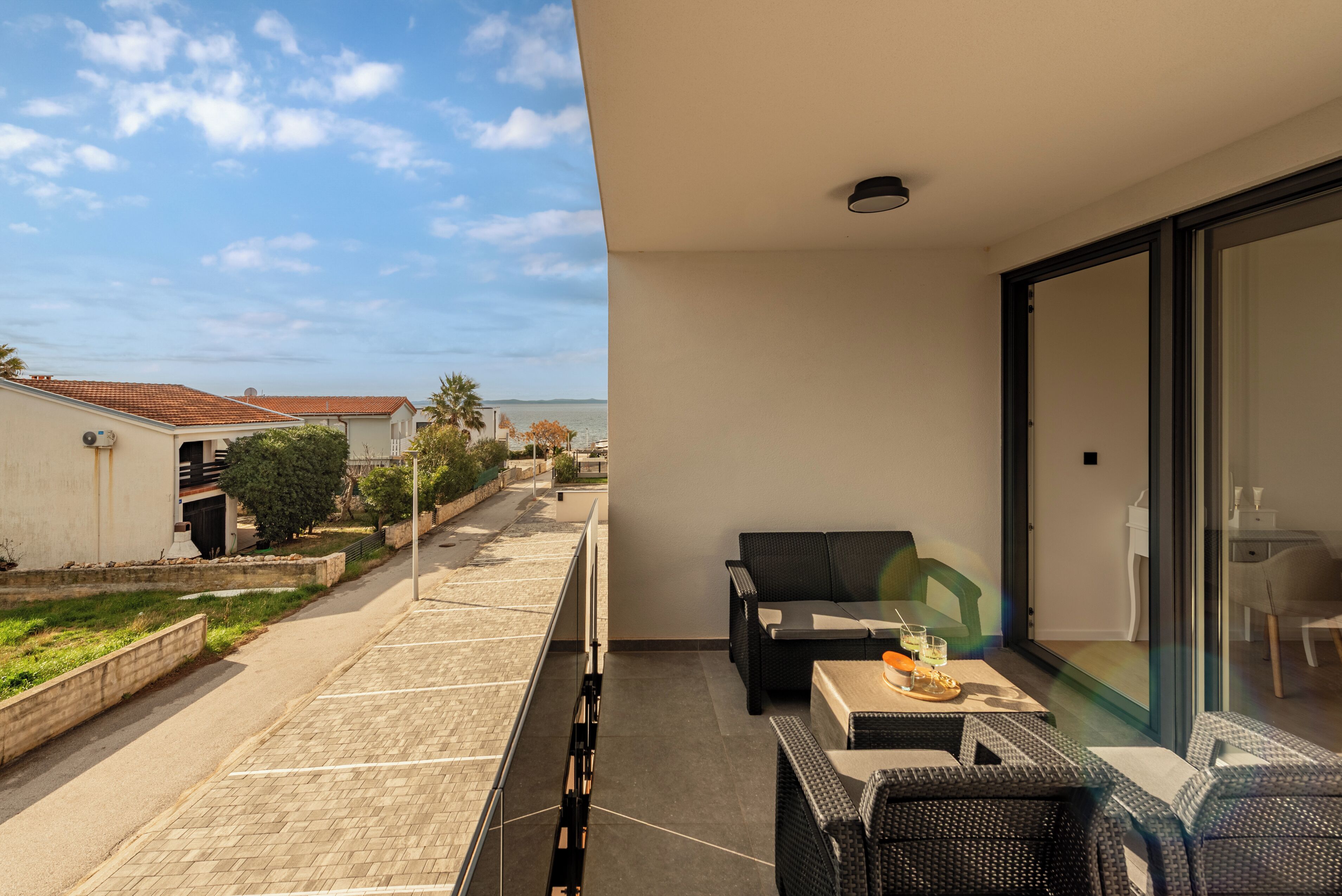 Ein Balkon mit Blick auf die Straße und das Meer, ein Tisch mit Stühlen und eine Schiebeglastür.
