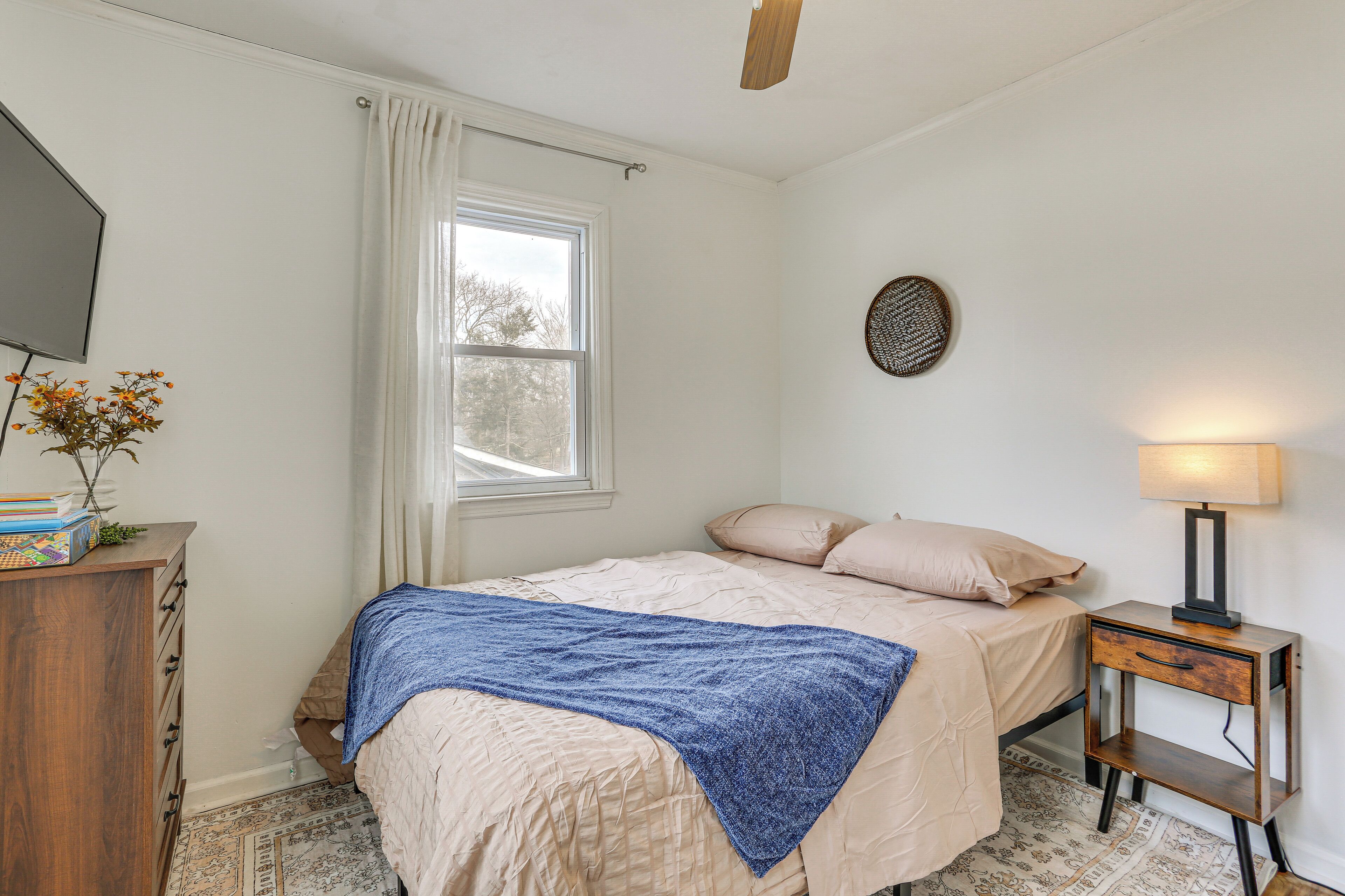 A bedroom with a bed, a nightstand with a lamp, a dresser, a window with curtains, and a wall-mounted decorative item.