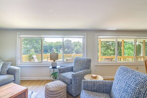 A living room with a view of a wooden deck and trees. There are two sofas, a coffee table, and a side table with a lamp.