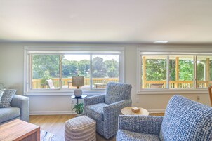 Sala de estar con vistas a una terraza de madera y árboles. Hay dos sofás, una mesa de centro y una mesita de noche con una lámpara.