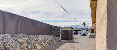 A gravel driveway leading to a gated area with a chain-link fence, a small utility shed, and a building with a stucco exterior.