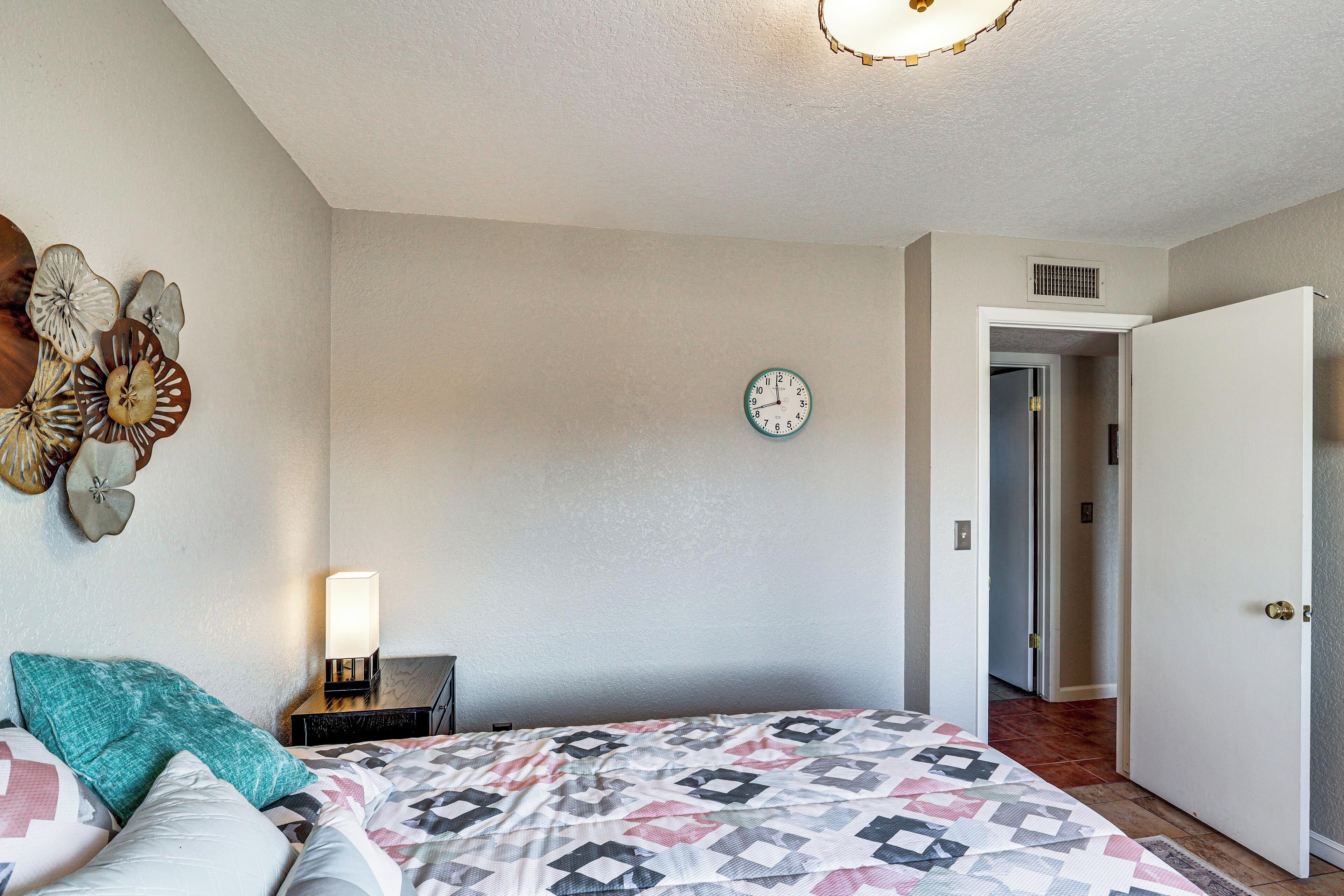 Quarto com cama, travesseiros, relógio na parede e porta que dá para outro cômodo.