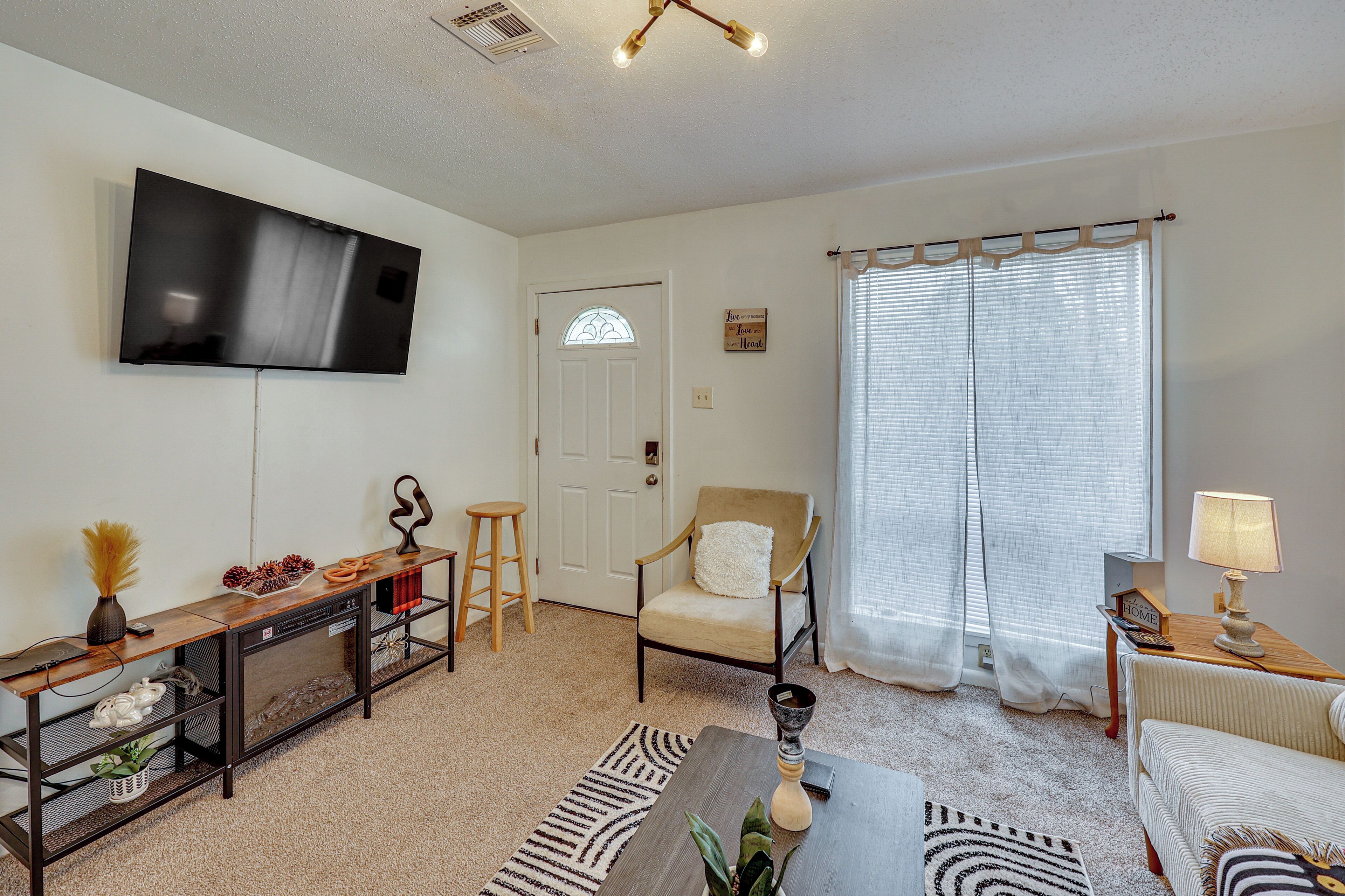 A living room with a flat-screen TV, a wooden coffee table, a beige armchair, a white sofa, and a large window with sheer curtains.