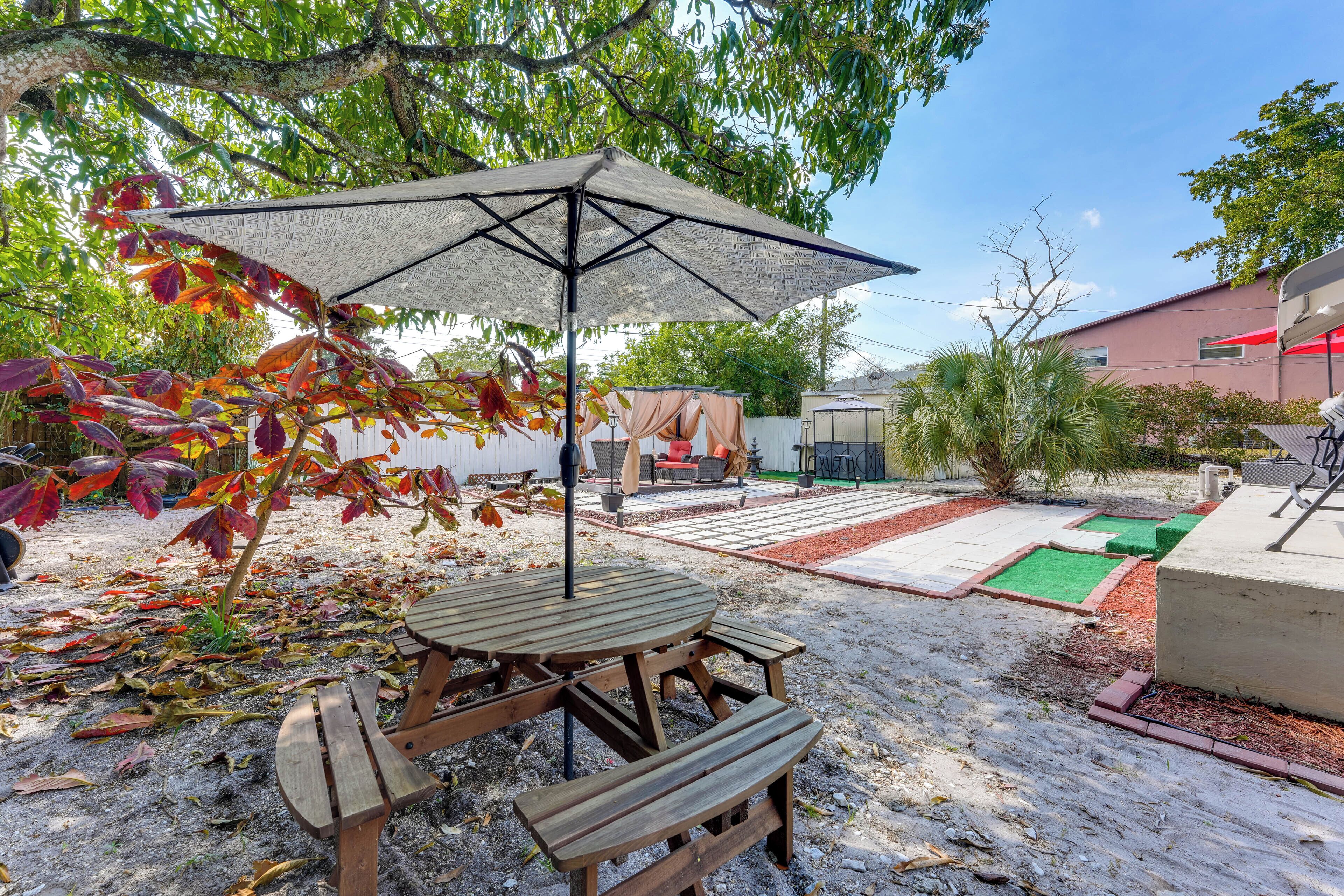 A wooden picnic table with a large umbrella, surrounded by a garden with various plants and a red building in the background.