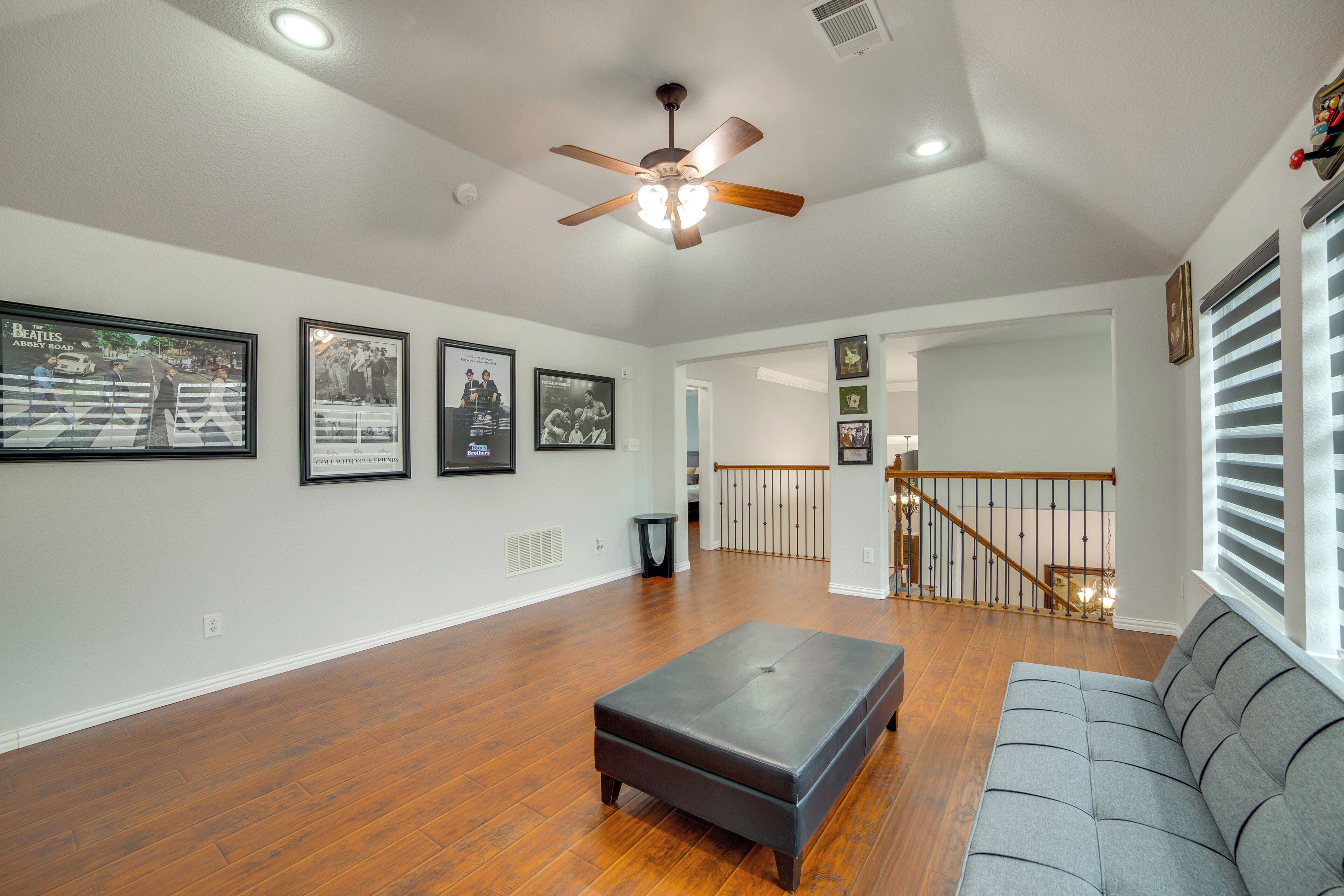 A spacious living room with hardwood flooring, a grey sofa, a black ottoman, and a ceiling fan.