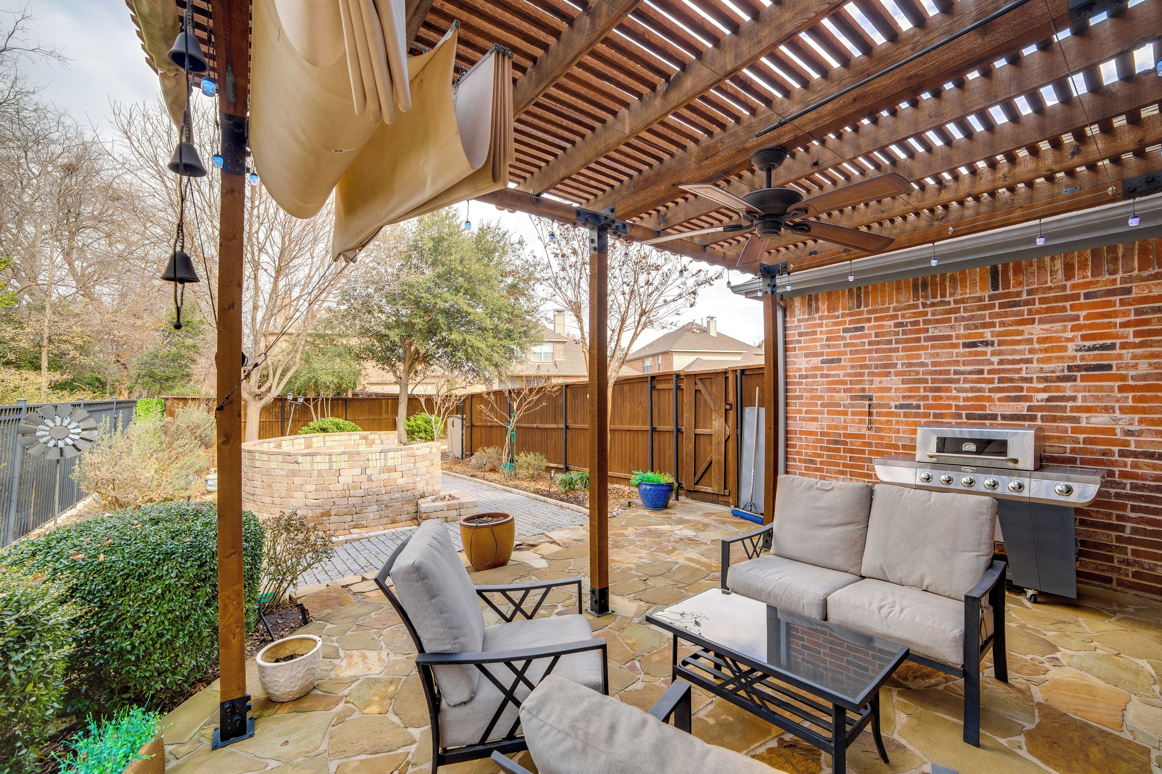 Un patio al aire libre con suelo de piedra, pared de ladrillo y una pérgola de madera. Se observa una parrilla y una zona de estar con sillas y una mesa.